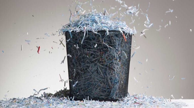 Shredded paper overflowing black mesh wastebasket.