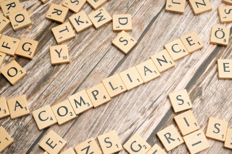 Scrabble tiles spelling 'Compliance' on wooden surface.